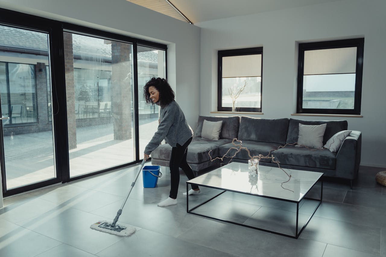 A woman mopping the floor in a bright, modern living room with large windows and stylish furniture.
